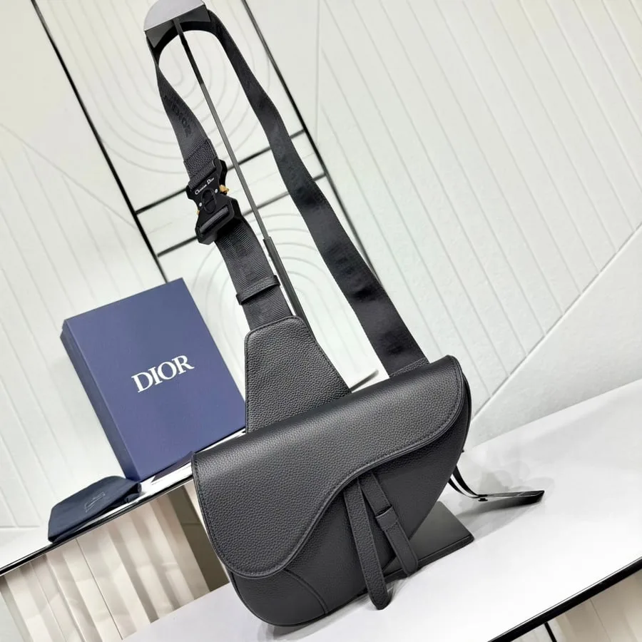 Black grained leather Saddle bag with its long strap coiled beside a Dior box, photographed at a three-quarter angle