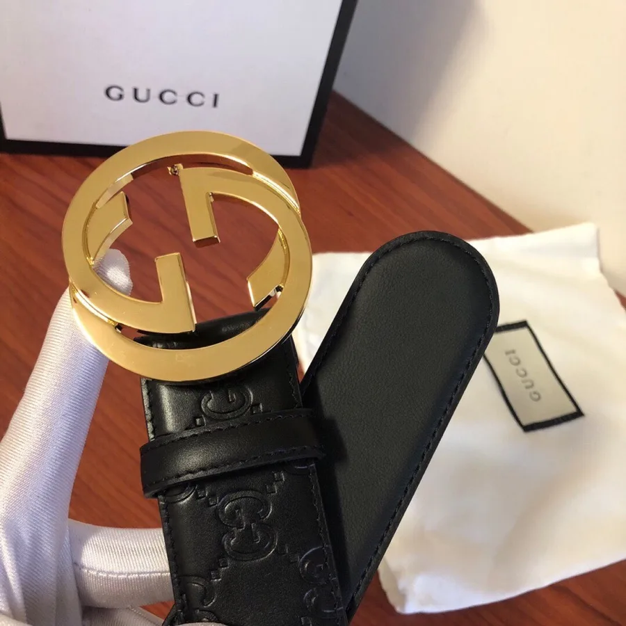 Gucci black leather belt with a gold-tone interlocking G buckle, embossed GG pattern along the strap and a dust bag behind, from the GunongTrade catalog