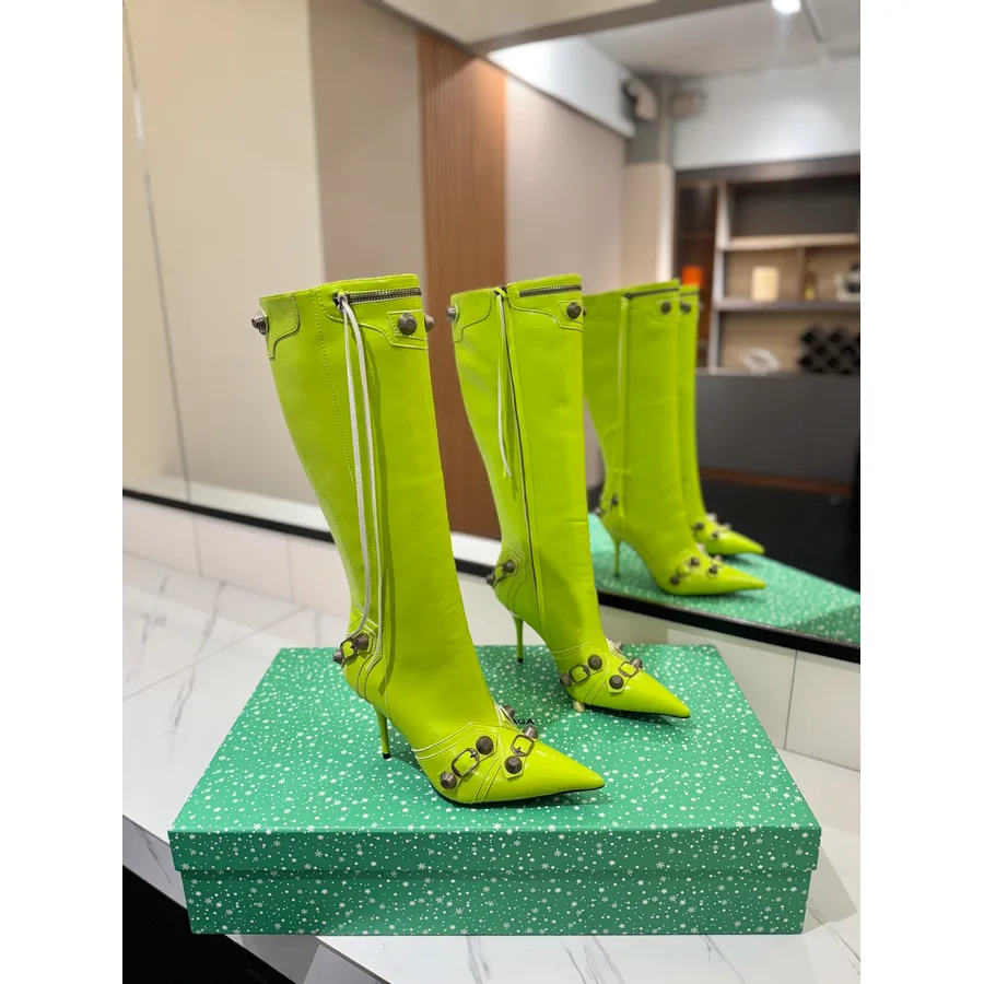 Bright green knee-high pointed-toe boot with silver studs and buckled straps on the shaft, a front zip and a thin stiletto heel