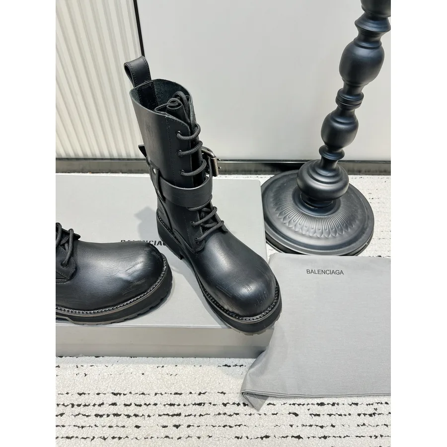 Black mid-calf lace-up boot with a buckled strap, a pull tab at the back and a chunky lugged rubber sole