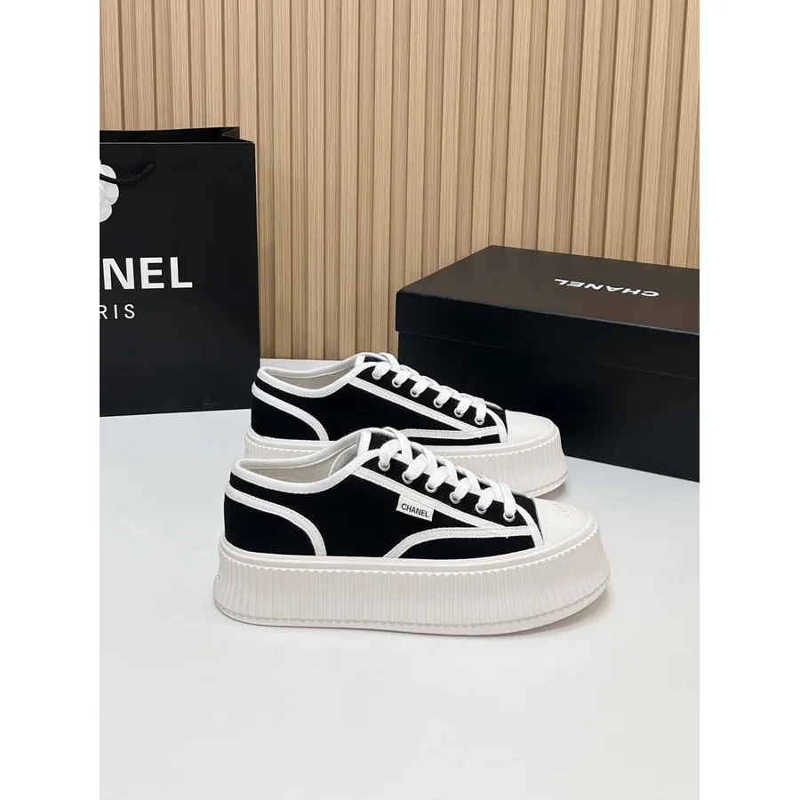 Black canvas Chanel sneaker shown side-on, with white piping and a white heel tab, white laces through white eyelets, a small rectangular Chanel logo tag on the quarter and a deep white ribbed platform sole