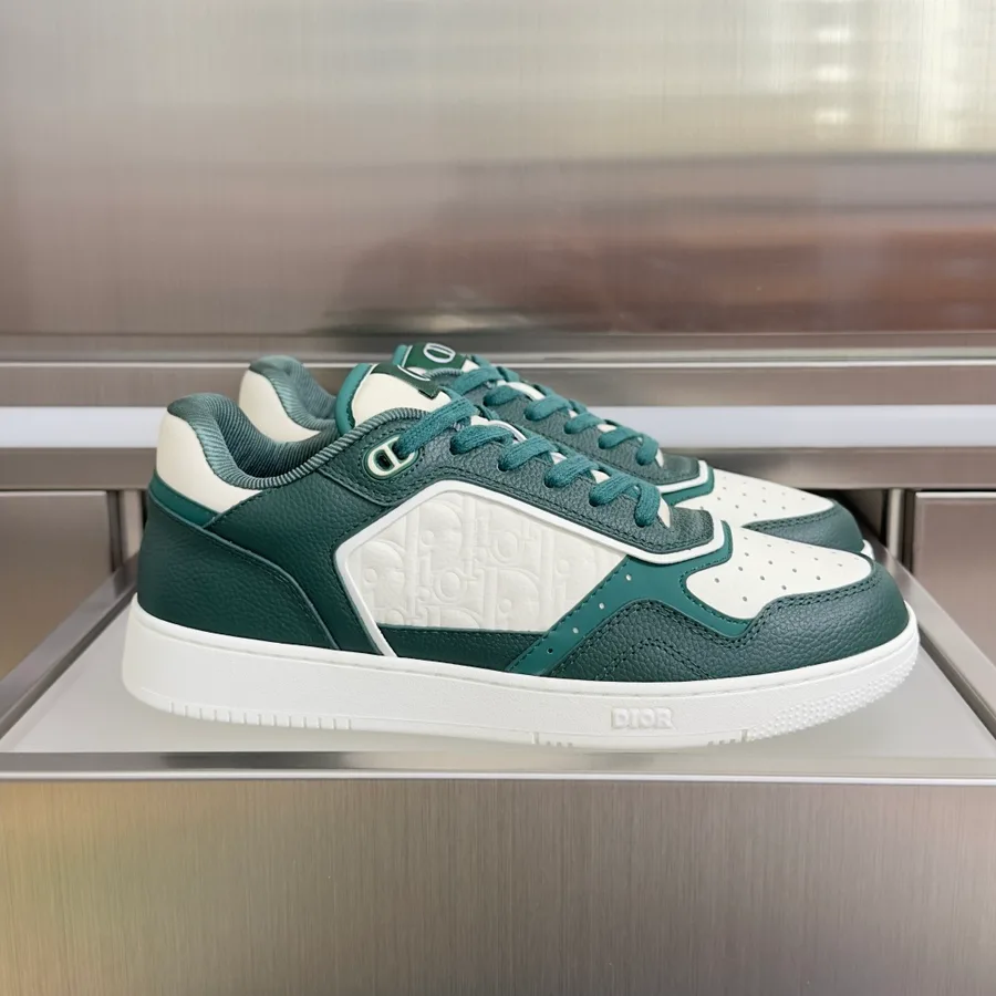 Green and white Dior B27 low-top sneaker photographed side-on, showing the round lace eyelets, the perforated toe box, an embossed Oblique panel across the quarter and the DIOR lettering moulded into the white rubber sole