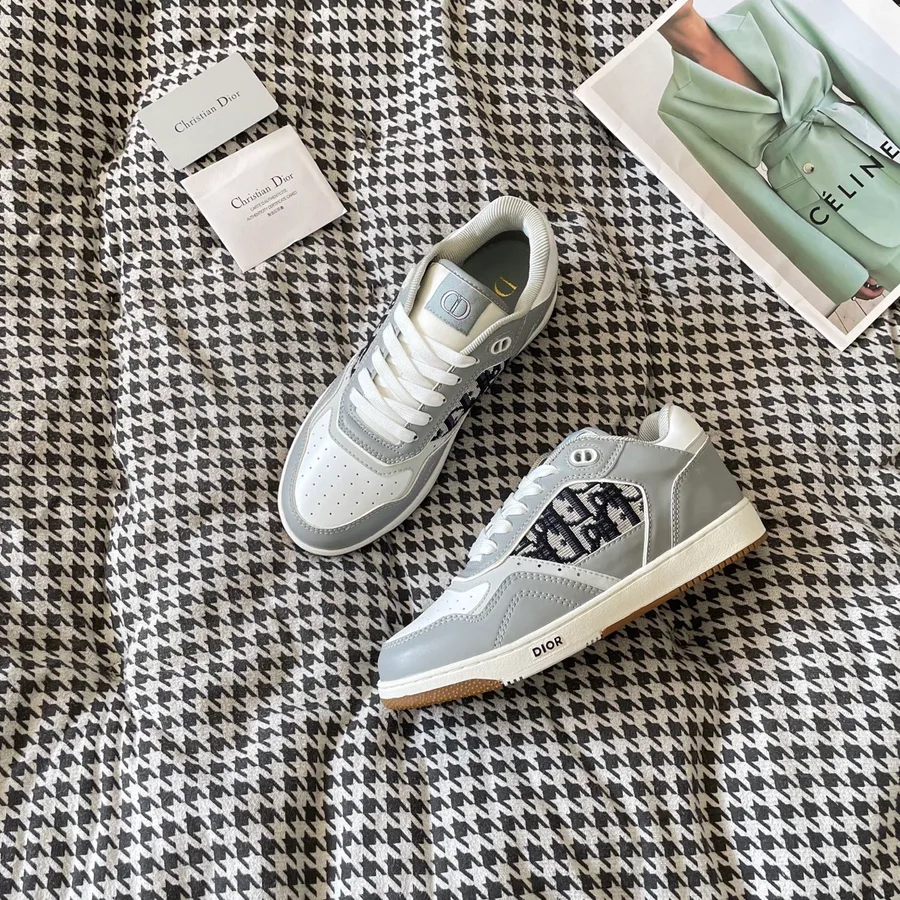 Grey and white Dior B27 low-top sneaker shown at a three-quarter angle, with a monogram-patterned side panel, white laces, grey leather overlays and the DIOR signature on the side of the sole