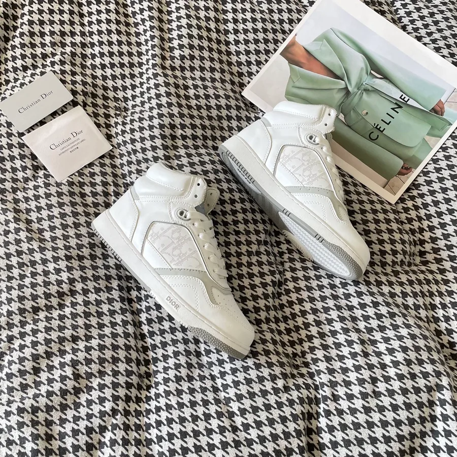 White Dior B27 high-top sneaker from the side, showing the taller collar above the ankle, the lace-up closure and the DIOR lettering on the side of the two-tone rubber sole