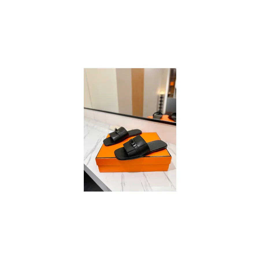 Black slide sandals with a wide strap fastened by a metal buckle, photographed on an orange box