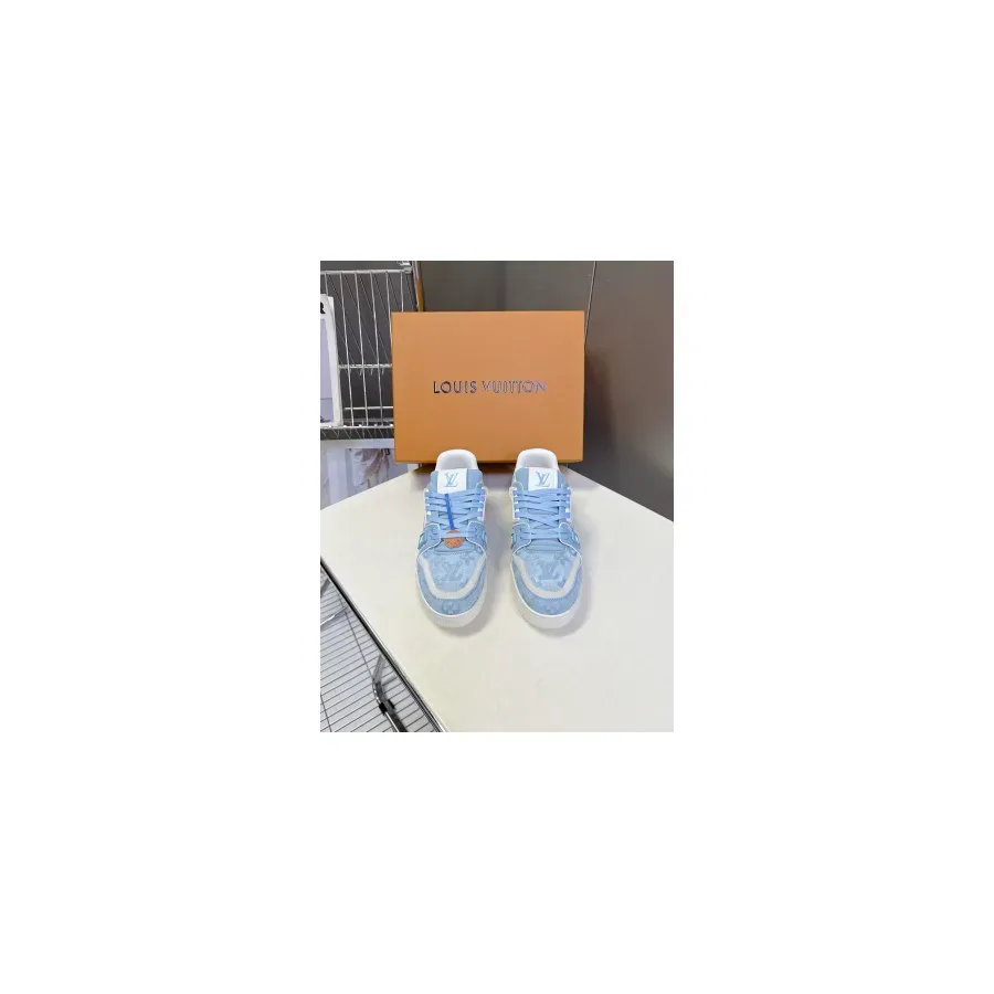 Pair of light blue LV Trainer low-top sneakers with thick white rubber soles, photographed in front of an orange Louis Vuitton box