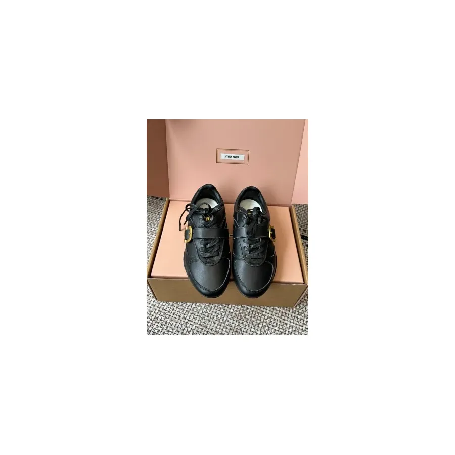 Black leather low-top sneaker with a lace-up front, a strap across the instep and a gold-tone buckle, shown beside a pink box with a label
