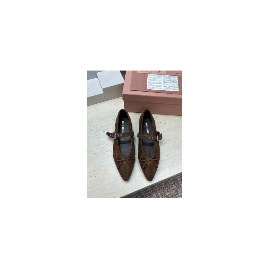 Brown suede pointed-toe flat with a bow at the front and a buckled strap across the instep, shown on a pink box