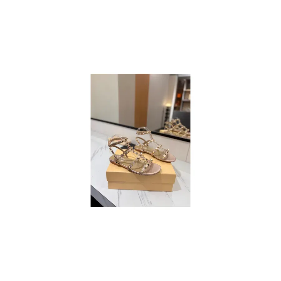 Nude studded T-strap sandal with an open toe, a flat sole, an ankle strap with a buckle and gold-tone studs along the straps
